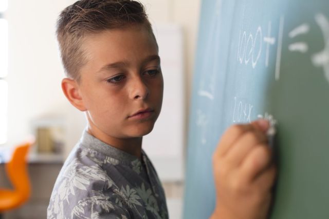 Rapaz em pé junto ao quadro-negro a escrever numa sala de aula durante a aula de matemática