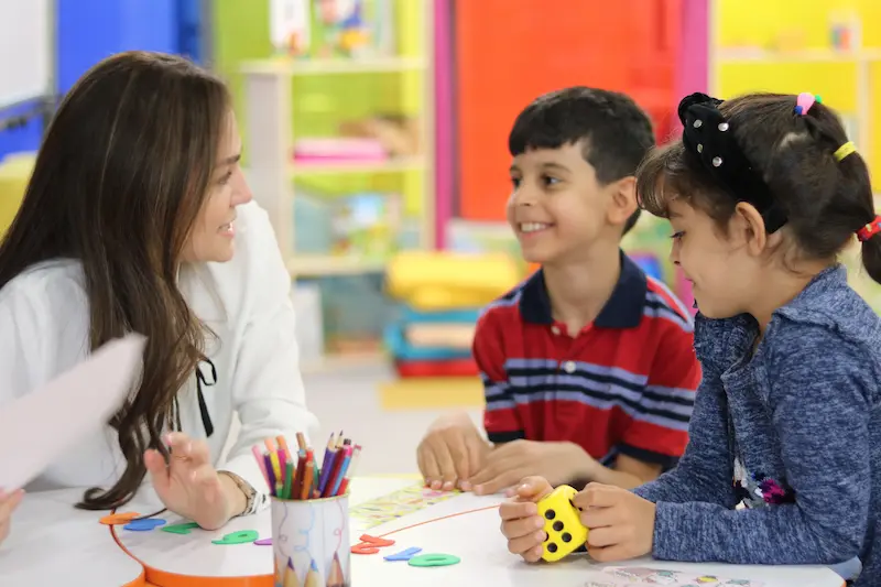Professora a apoiar duas crianças em atividades práticas de aprendizagem visual, num ambiente de sala de aula inclusivo e colorido